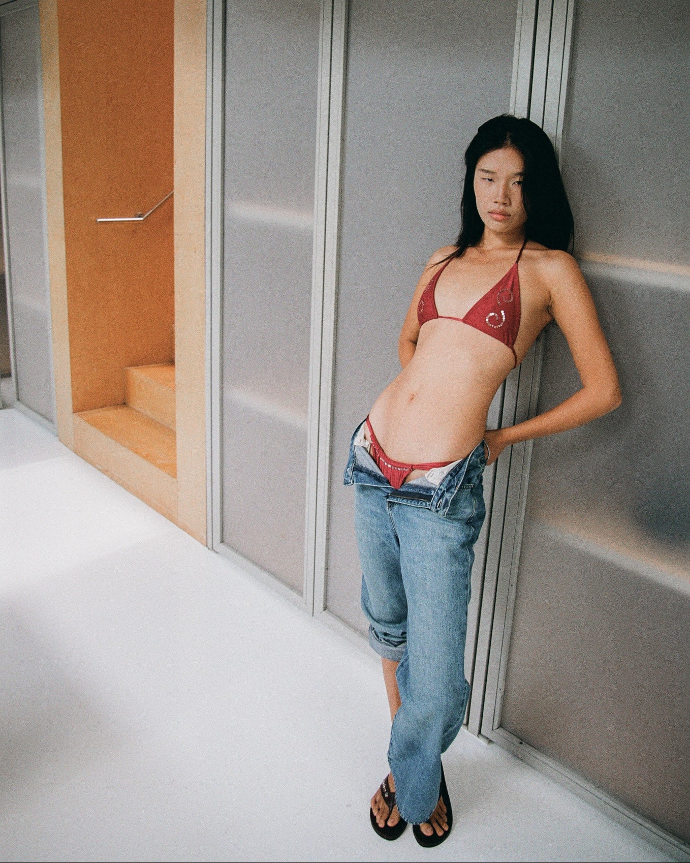Woman wearing a maroon spiral sequin bikini top and blue jeans standing against a metallic wall.