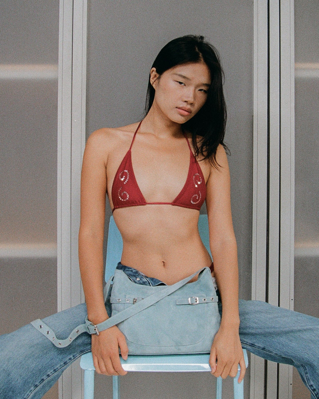 Woman wearing a maroon bikini top and blue denim jeans, holding a light blue suede bag against a frosted glass background.