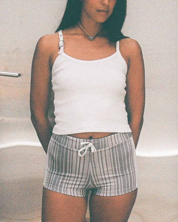 Woman standing in front of a silver door with concrete walls and floor wearing a white tank top and striped grey shorts. 